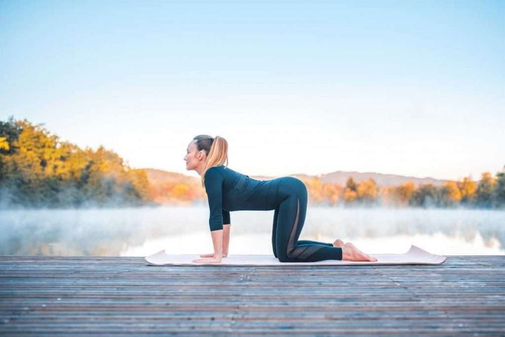 Una mujer haciendo pose de vaca en una alfombra de yoga afuera.